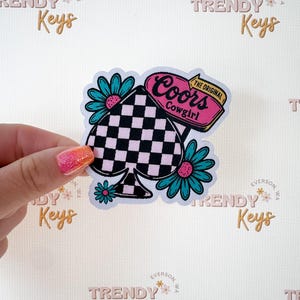 May include: A sticker featuring a black and white checkered spade shape with the text "Coors Cowgirl" in pink. Teal flowers with pink centers surround the spade. The sticker is held by a hand with pink and orange glitter nail polish.