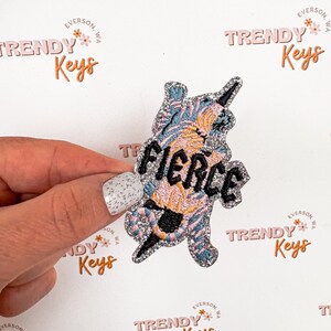 May include: A sparkly blue and pink tiger with a silver glitter outline and the word "FIERCE" in silver glitter letters. The tiger is holding a silver glitter lightning bolt. The image is on a white background with the logo "TRENDY Keys" repeated in the background.