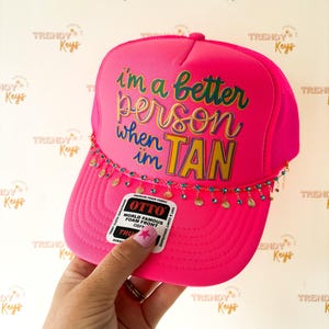 May include: A bright neon pink trucker hat featuring the text "i'm a better person when im TAN" in multi-colored lettering. The hat is adorned with a delicate gold-tone chain fringe featuring small turquoise and gold circular charms.