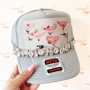 May include: A light gray trucker hat featuring a graphic print of four pink cocktails in martini glasses. The hat is embellished with a row of clear, faceted rhinestones across the front panel and includes an Otto brand sticker on the brim.