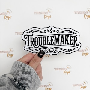 May include: A black and white patch with the word "TROUBLEMAKER" in bold lettering. The patch has a decorative border and star accents. The patch is held by a person with a gray sleeve and black nail polish.