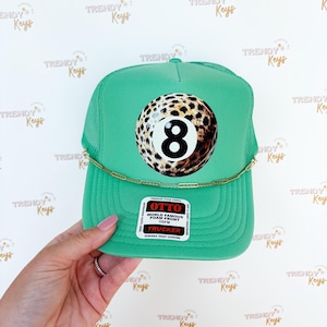 May include: A green trucker hat with a mesh back and a curved brim. The front features a large graphic of an eight ball with a leopard print design. The hat has a gold chain detail and a tag that reads "OTTO TRUCKER".