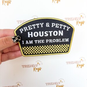May include: Black and yellow patch with the text "PRETTY & PETTY HOUSTON I AM THE PROBLEM". The patch has a black background with white text and a yellow border. A checkered pattern is at the bottom.