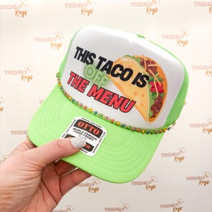 May include: A green and white trucker hat with a colorful rhinestone trim. The hat features a graphic of a taco with the text "This Taco Is Off The Menu".