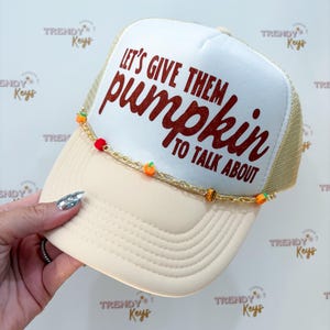 May include: A cream-colored trucker hat with a white front panel featuring the glittery dark red text "LET'S GIVE THEM pumpkin TO TALK ABOUT". A gold-tone chain is draped across the brim, accented with small orange pumpkin and red bead charms.