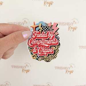 May include: Embroidered patch with the text "Fueled by Compliments & Chaos" in red script. The patch features a mix of designs, including a lightning bolt, stars, and a checkered flag, with a leopard print and smiley faces.