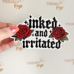 May include: Embroidered patch with the words "inked and irritated" in black gothic lettering. Two red roses with green leaves flank the text. The patch is white with a black border.