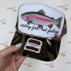 May include: A trucker hat with a white front panel featuring a colorful fish illustration and the text "catching fish, not feelings." The hat has a camouflage pattern on the brim and a gold chain detail.