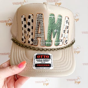 May include: Tan trucker hat with the word "MAMA" in large, textured letters. The "M" is patterned with soccer balls, the "A" with a net design, the second "M" with green soccer field texture, and the final "A" with soccer jerseys. A chain detail accents the brim.