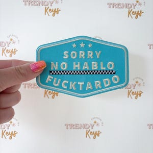 May include: A turquoise embroidered patch with the words "SORRY NO HABLO FUCKTARDO" in white lettering. The patch has a black and white checkered stripe and three stars at the top. The background is white with a repeating logo.