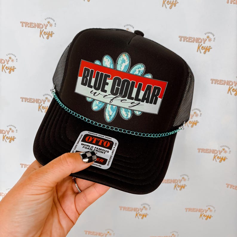 Puede incluir: Gorra de camionero negra con parte trasera de malla y visera curva. La parte delantera presenta el texto "BLUE COLLAR wifey" en blanco, rojo y azul, con un dise&ntilde;o floral. Una cadena azul claro adorna la visera.