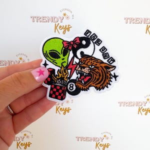 May include: Embroidered patch featuring a green alien, a yin-yang symbol with a red bow, and a tiger's head. A red lightning bolt and a checkered lighter with the number 8 are also included. The text "FAR OUT" is above the tiger.