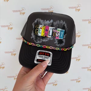 May include: A black trucker hat with a mesh back. The front of the hat features a gray and white graphic of trees and license plates that spell out "GET LOST". The hat has a colorful chain trim around the brim.