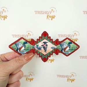 May include: An embroidered patch with a central image of ducks in a pond, flanked by duck images. The patch has a red border with a green and red geometric design. The text "TRENDY Keys" is visible in the background.