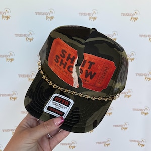 May include: A camouflage trucker hat with a red and white ticket design that says "Shit Show" and "Keep this cool on". The hat has a gold chain around the brim.