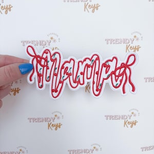 May include: A white patch with the word "Mama" in red, cursive lettering. The letters are outlined in red and filled with small green and red cherries. The patch is held against a white background with the text "Trendy Keys" repeated.