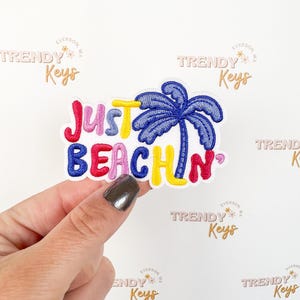 May include: Embroidered patch with the words "Just Beachin" in colorful letters, featuring a blue palm tree. The patch is held against a white background with the words "Trendy Keys" repeated.