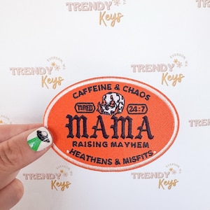 May include: An orange embroidered patch with the text "MAMA" in black, surrounded by the phrases "Caffeine & Chaos," "Raising Mayhem," and "Heathens & Misfits." A skull graphic is in the center, with "Tired" and "24:7" on either side.