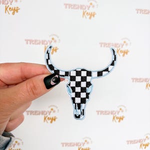 May include: A black and white checkered bull skull patch with light blue trim. The patch is an embroidered design with a skull shape and curved horns. The background is white with the words "TRENDY Keys" repeated.