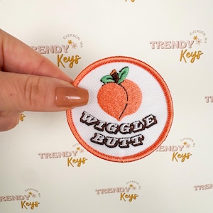 May include: Embroidered patch featuring a peach design with the text "Wiggle Butt" in brown letters. The patch has a white background with an orange border and a green leaf.