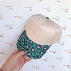 May include: A beige baseball cap with a green and pink leopard print brim. The cap has a green button on top. The background has a repeating pattern of the words "TRENDY Keys".