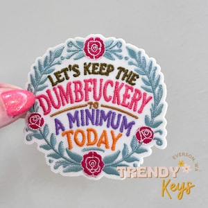 May include: Embroidered patch with the text "LET'S KEEP THE DUMBFUCKER TO A MINIMUM TODAY" in pink, orange, and purple. The patch is surrounded by a wreath of blue leaves and pink roses. The patch is held by a hand with pink nail polish.