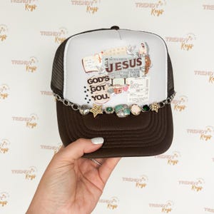 May include: A brown and white trucker hat with a mesh back. The front of the hat has a collage of images and text, including the words "God's Got You" and "Do You Know Jesus". The hat is decorated with a silver chain and charms.