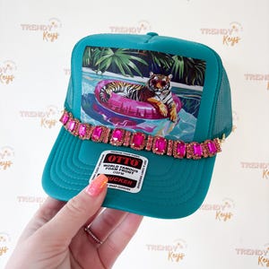 May include: Teal trucker hat with a vibrant image of a tiger in a pool float. The hat features a pink jeweled band across the brim and the text "OTTO WORLD FAMOUS FOAM FRONT".