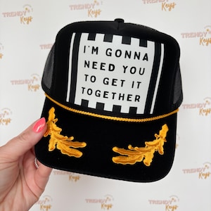 May include: Black trucker hat with a mesh back and a rectangular patch that reads "I'M GONNA NEED YOU TO GET IT TOGETHER" in white block letters. The hat features gold embroidered leaf accents and a gold rope trim.