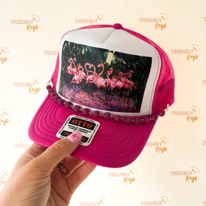 May include: A bright pink trucker hat featuring a white foam front panel with a graphic print of a flock of pink flamingos. The hat includes a pink mesh back, a pink brim, and a decorative string of small, sparkling pink beads across the base.