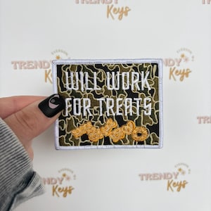May include: Square patch with a camouflage background and the words "WILL WORK FOR TREATS" embroidered in white. The patch has a white border and a gold-colored design at the bottom.