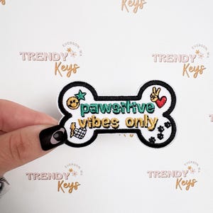 May include: A white, bone-shaped patch with the words "pawsitive vibes only" in green and gold embroidery. The patch features a smiley face, peace sign, heart, and other small graphics. The patch is held by a hand.