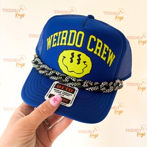 May include: A royal blue trucker hat featuring the yellow text WEIRDO CREW above a yellow distorted smiley face graphic. A black and white checkered chain is attached across the front panel, and an Otto brand tag is affixed to the brim.