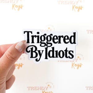 May include: A white patch with the words "Triggered By Idiots" in black, stylized lettering. The patch is rectangular with a white border and is held by a hand against a white background with the words "Trendy Keys" in a repeating pattern.