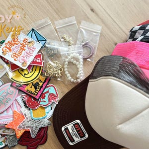 May include: A collection of colorful embroidered patches with various designs, including a smiley face, a pumpkin, and a mermaid. The patches are arranged on a brown surface with a brown and white trucker hat, a pink and black trucker hat, and a black and white checkered trucker hat.