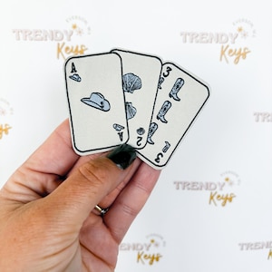 May include: Three playing card patches are held in a hand. The cards feature illustrations of a cowboy hat, seashells, and cowboy boots. The cards are cream-colored with black outlines and details. The background has the words "TRENDY Keys" repeated.