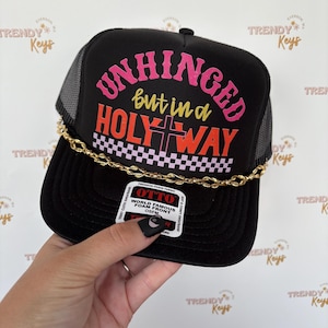 May include: Black trucker hat with the text "UNHINGED but in a HOLY WAY" in pink, yellow, and orange. The hat has a gold chain detail and a mesh back. The hat is being held by a person.