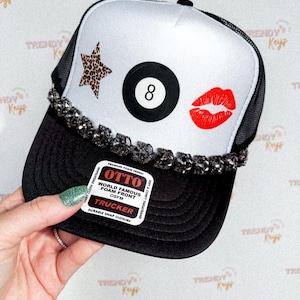 May include: Black and white trucker hat with a white front panel featuring a star, an eight ball, and red lips. The hat has a black brim and mesh back, and a decorative jeweled band.