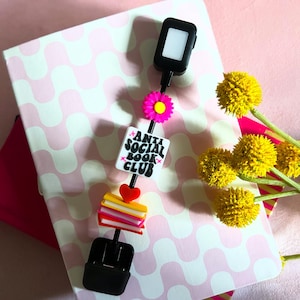 May include: A black and white bookmark with the text "Anti Social Book Club" in a retro font. The bookmark is attached to a black chain with a pink flower charm, a stack of colorful books charm, and a black phone charm.