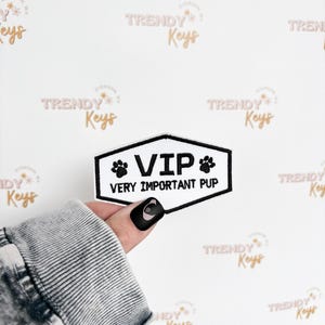 May include: A black and white patch with the text "VIP VERY IMPORTANT PUP" and paw prints. The patch is held against a white background with the words "TRENDY Keys" in a repeating pattern.