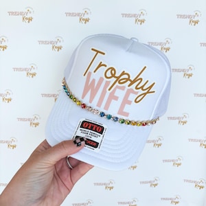 May include: White trucker hat with a pink and gold "Trophy Wife" design. The hat has a rainbow-colored rhinestone trim.