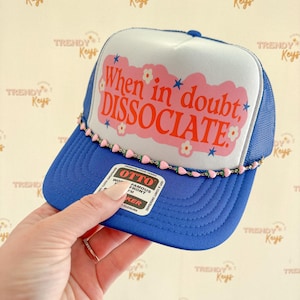 May include: A blue and white trucker hat with the text "When in doubt, DISSOCIATE" in pink. The hat has a mesh back and a curved brim. A pink and green beaded trim accents the brim. The hat is being held by a hand.