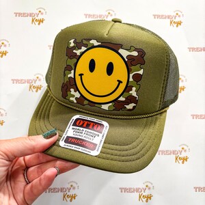 May include: An olive green trucker hat featuring a yellow smiley face graphic centered on a camouflage square background. The hat includes a foam front, a mesh back, and a sticker on the brim that reads OTTO World Famous Foam Front.