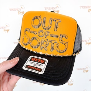 May include: Black and yellow trucker hat with the text "OUT OF SORTS" in black. The hat has a black brim and mesh back. A gold star-shaped trim accents the front panel. The hat has a snap closure.