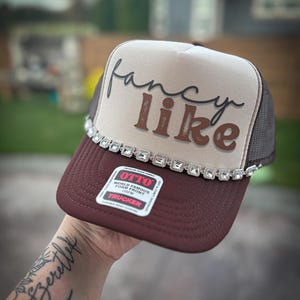 May include: A brown and beige trucker hat with a brown "fancy like" text on the front. The hat has a row of rhinestones around the brim.