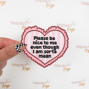 May include: A pink heart-shaped patch with a scalloped edge and the text "Please be nice to me even though I am sorta mean" in black. The patch has a vertical striped pattern in the center.