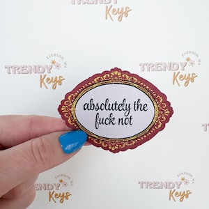 May include: An oval sticker with a burgundy and gold ornate border. The sticker has the words "absolutely the fuck not" in a black font on a white background. The sticker is held by a hand with a blue painted nail.