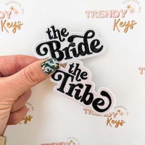 May include: Two embroidered patches with the phrases "the Bride" and "the Tribe" in black script lettering. The patches have a white background with a black border. The image also includes the text "Trendy Keys" and "Everson, WA".