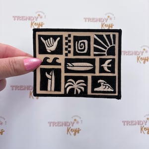May include: A rectangular embroidered patch featuring a grid of black and beige squares. The design includes icons of a shaka hand sign, a spiral, a sun, waves, a surfboard, a palm tree, and a crescent moon, all outlined in black thread.