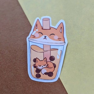 May include: A sticker of a cartoon fox sleeping inside a boba tea cup with tapioca pearls. The fox is orange and white with a brown nose and closed eyes. The boba tea is brown with a clear plastic cup and a straw.